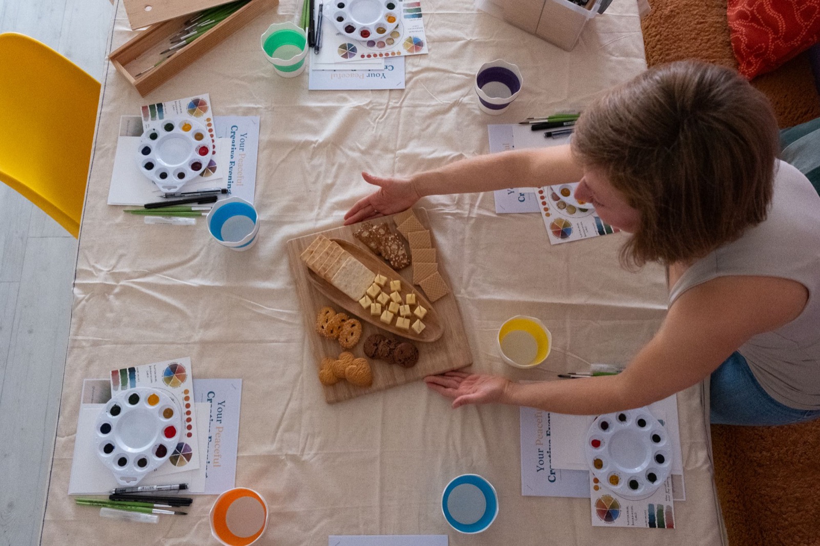 Gabriela setting a snack board on a workshop table laid with palettes, brushes, pens and colour cards