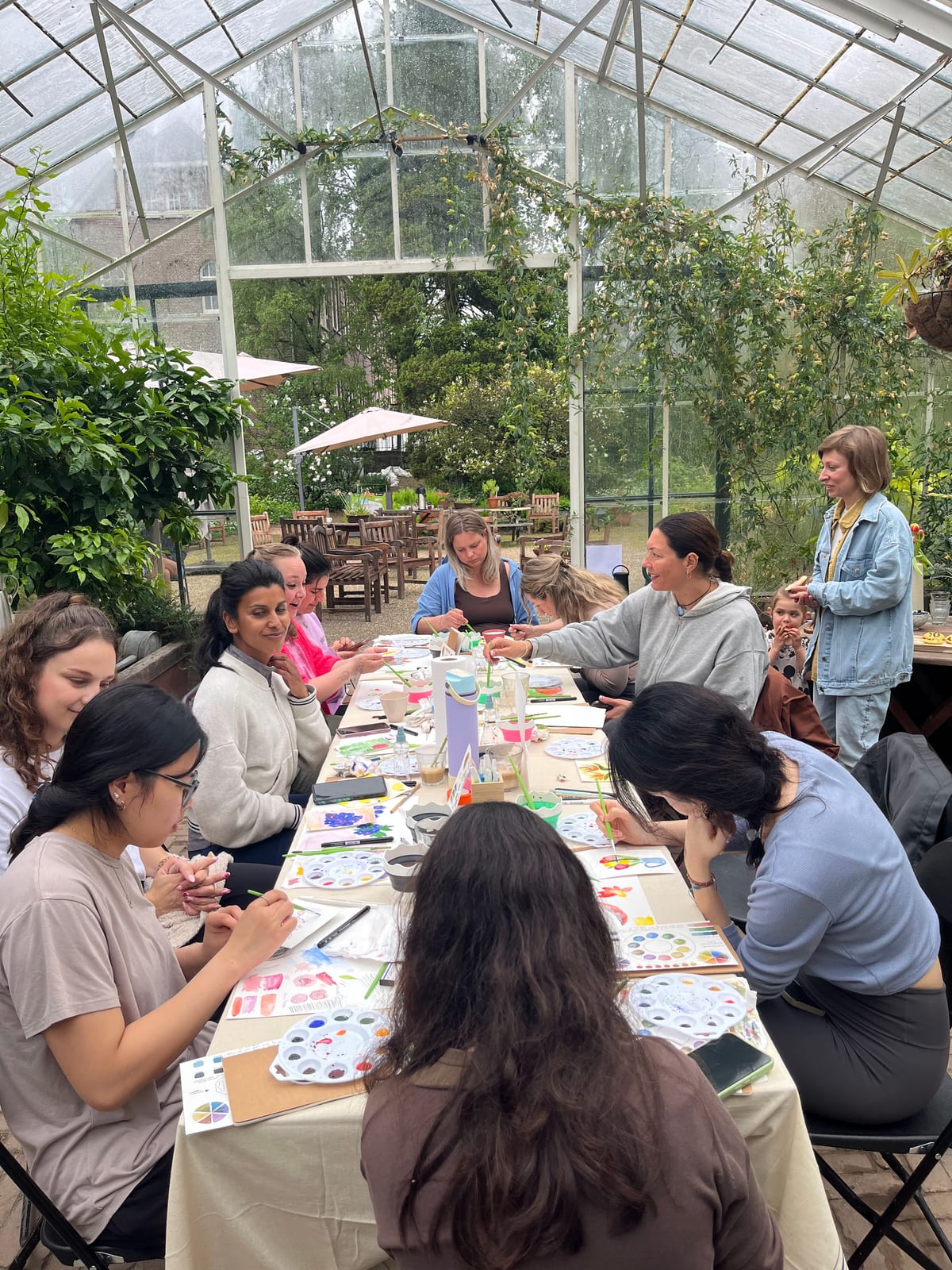 A group painting together at a long table inside a leafy greenhouse