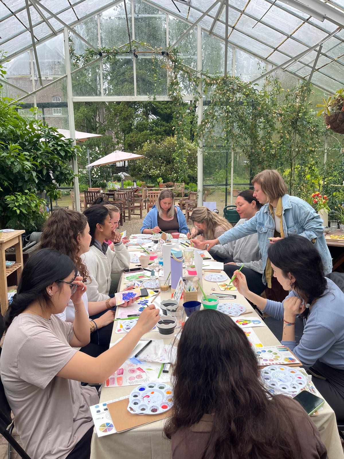 Gabriela leaning over a long table in a greenhouse, helping a group paint with watercolour