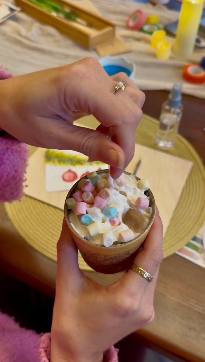 Hands adding pastel marshmallows to a hot chocolate, with a painted Christmas card behind